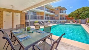 A poolside area with a dining table set for four, a covered seating area, and a two-story building with balconies in the background. - Copacabana Shores (Copacabana)