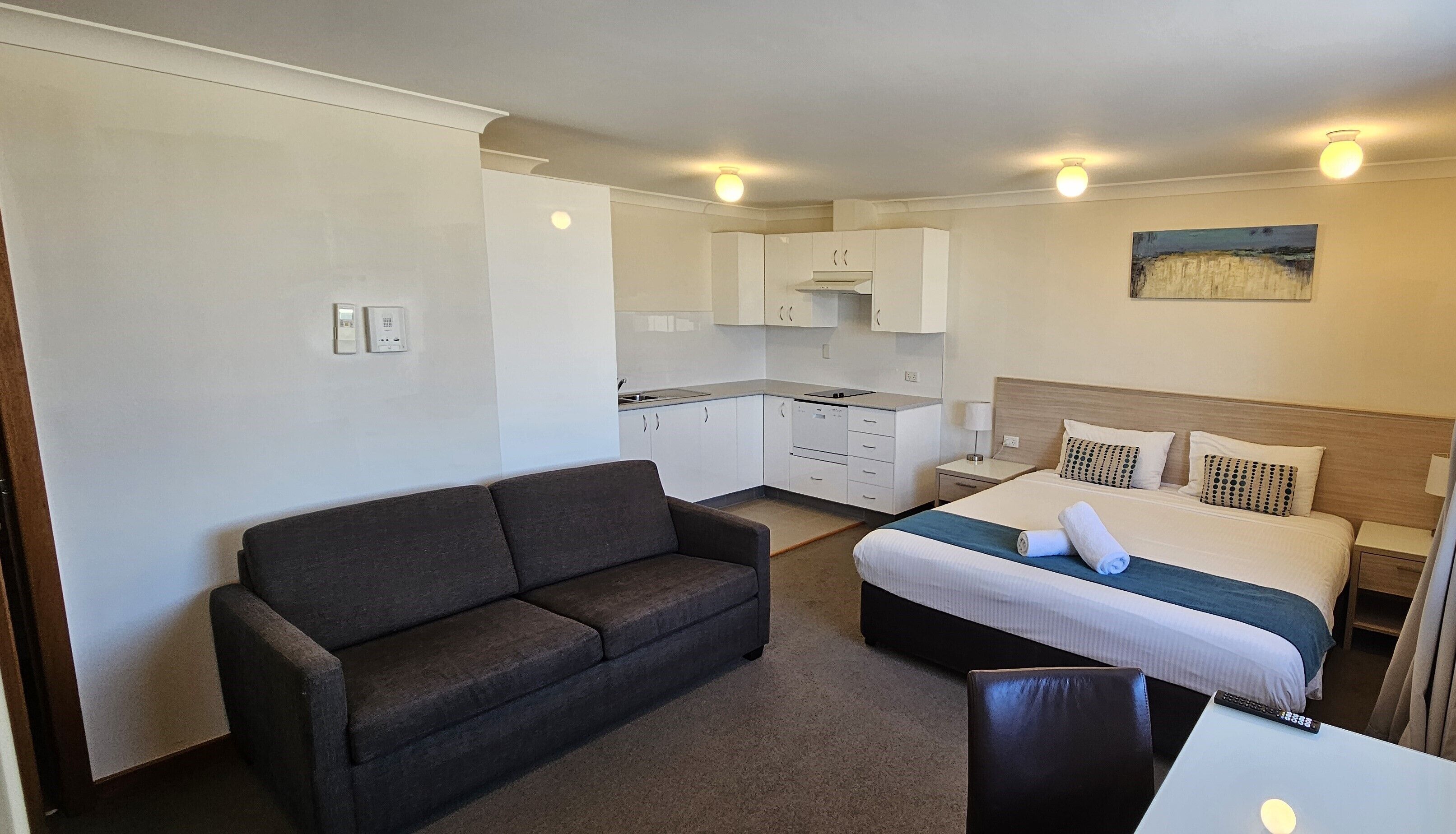 A compact hotel room with a bed, a sofa, a small kitchen area, and a painting on the wall.