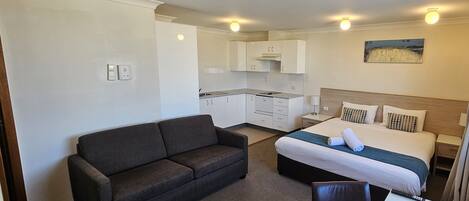 A compact hotel room with a bed, a sofa, a small kitchen area, and a painting on the wall.