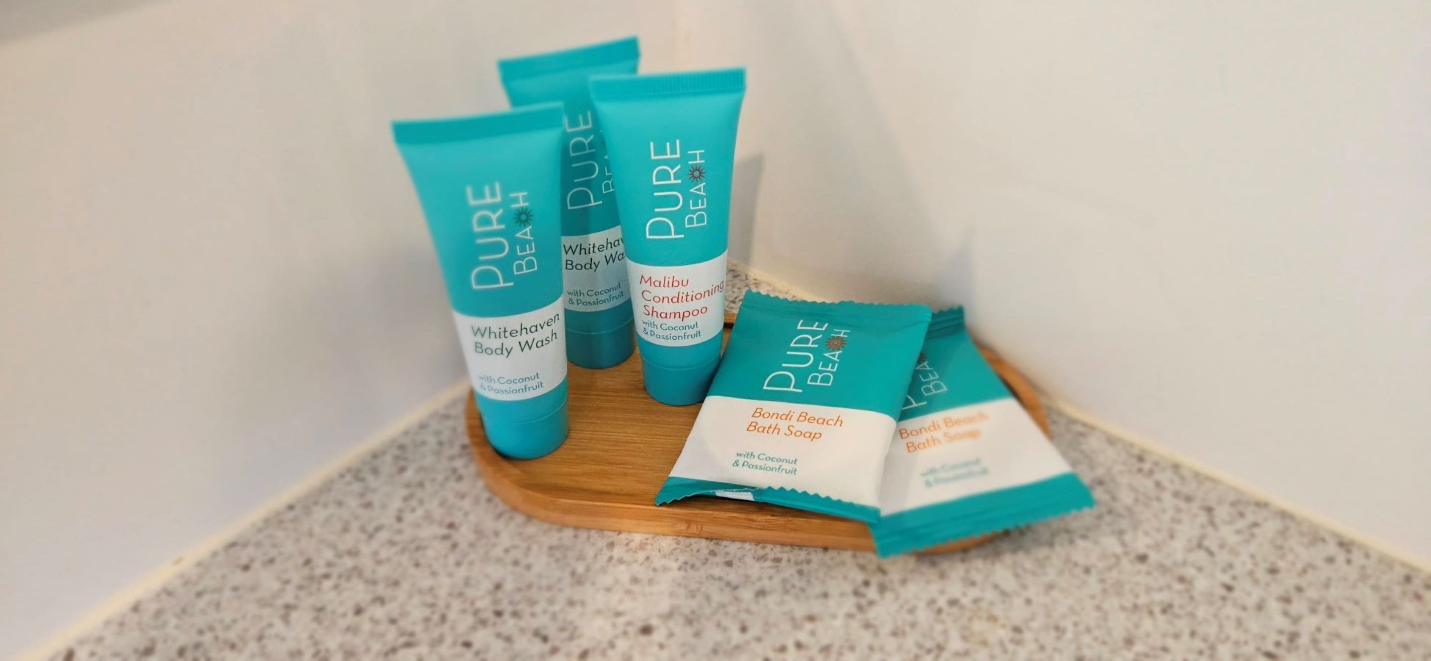 A set of Pure Beach body wash and shampoo tubes and bath soap packets on a wooden tray.