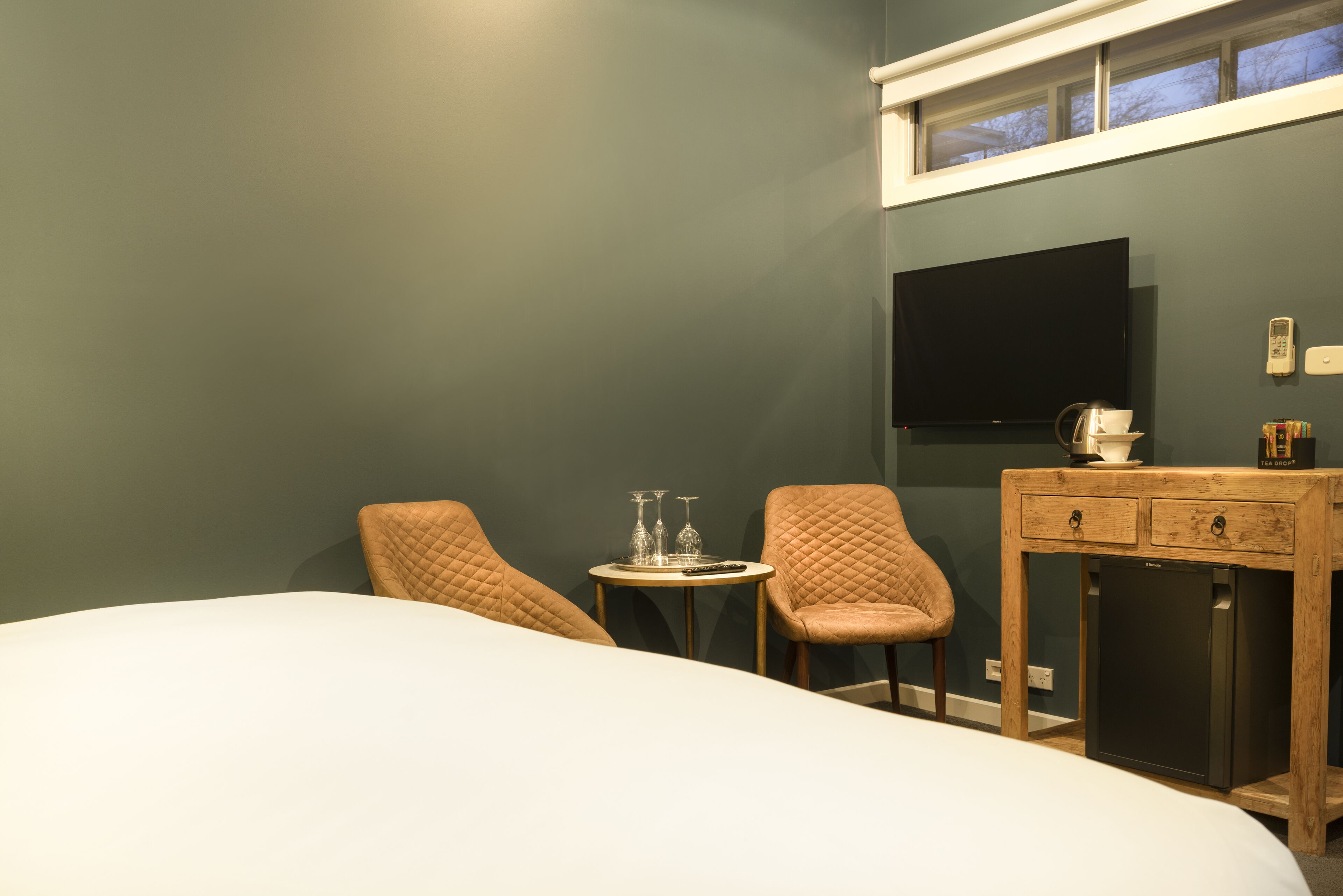 Habitación de hotel con cama, dos sillas, una mesita con vasos, cafetera y televisión en la pared.