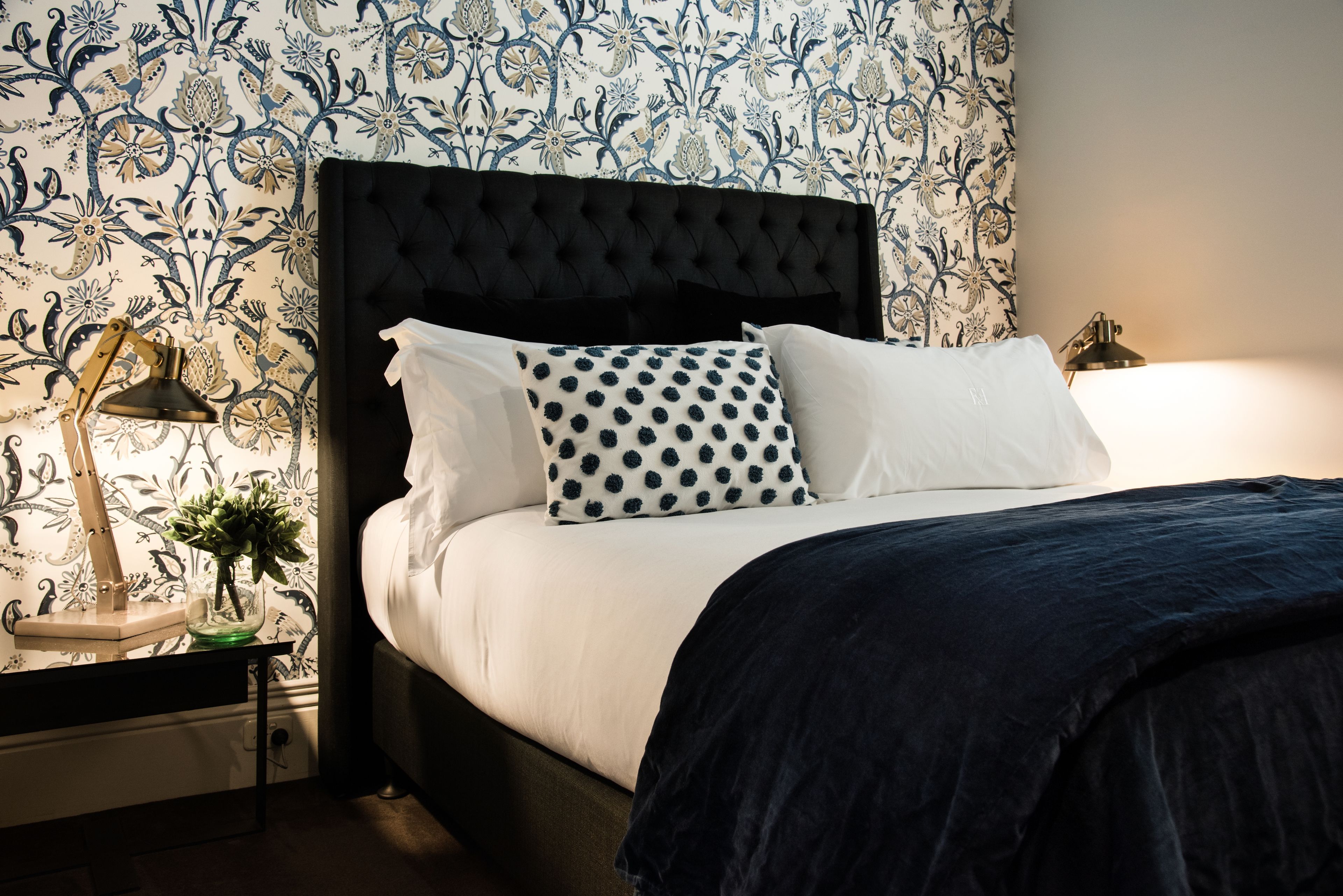 Cama bem arrumada com cabeceira preta, roupa de cama branca e almofada de bolinhas. Um abajur está sobre um criado-mudo com um vaso de flores.