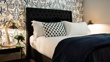 Cama bem arrumada com cabeceira preta, roupa de cama branca e almofada de bolinhas. Um abajur está sobre um criado-mudo com um vaso de flores.