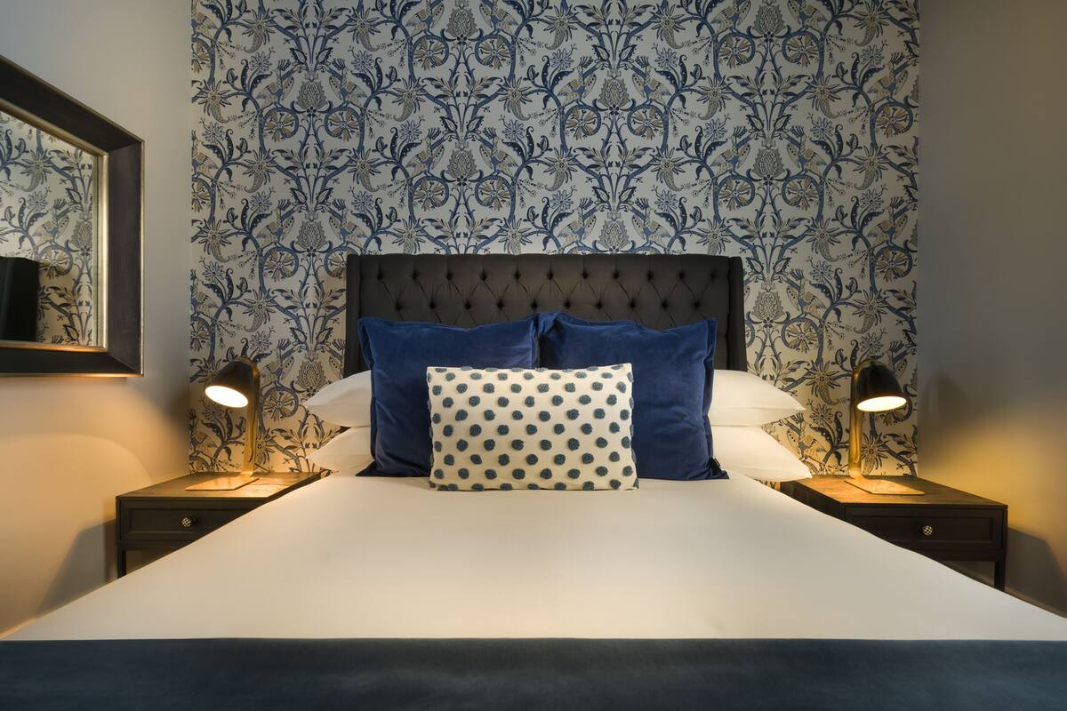 A neatly made bed with a tufted headboard, two bedside lamps, and a patterned wallpaper.