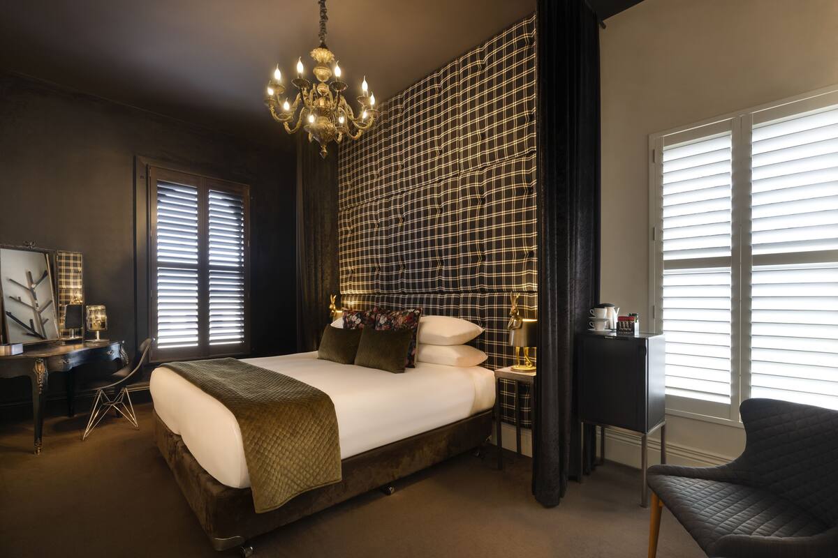 A bedroom with a large bed, a desk with a chair, a chandelier, and a view through the window.