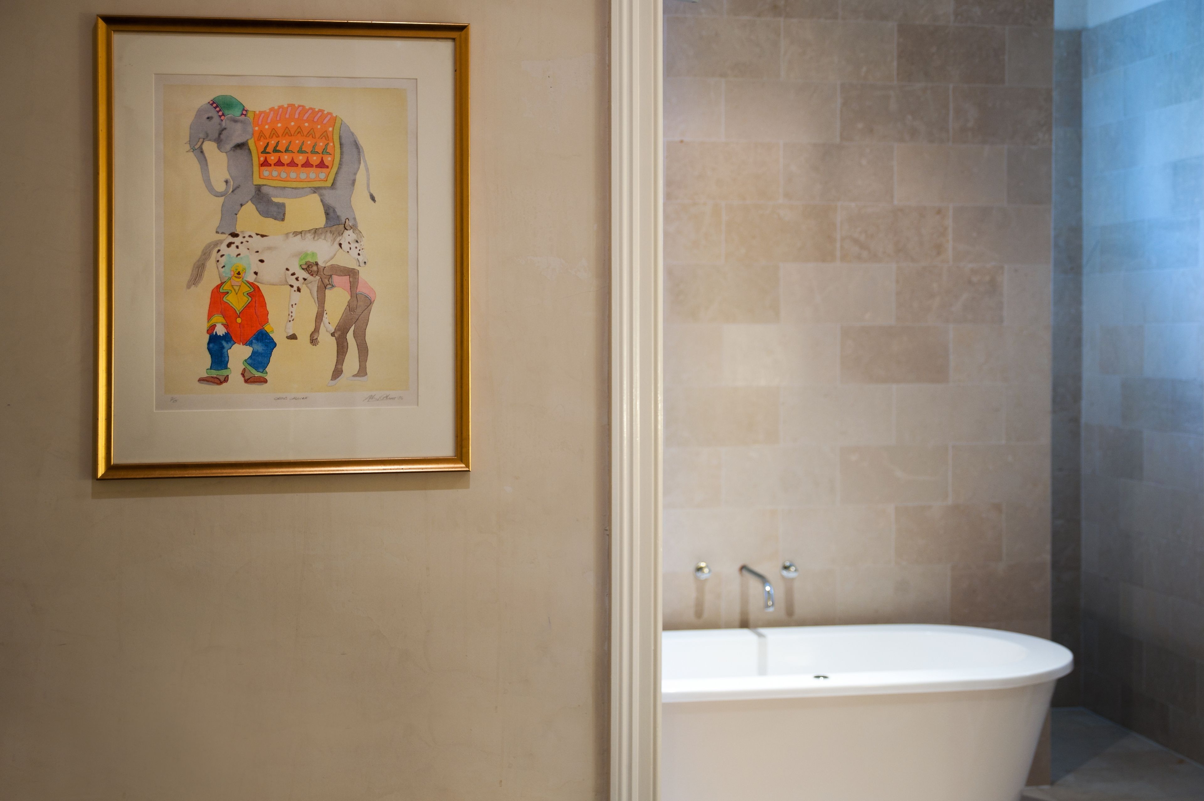 Une œuvre d’art encadrée représentant un éléphant et deux personnages, accrochée à un mur, une salle de bain avec des carreaux de couleur beige et une baignoire blanche.