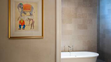A framed artwork of an elephant and two figures on a wall, a bathroom with beige tiles, and a white bathtub.