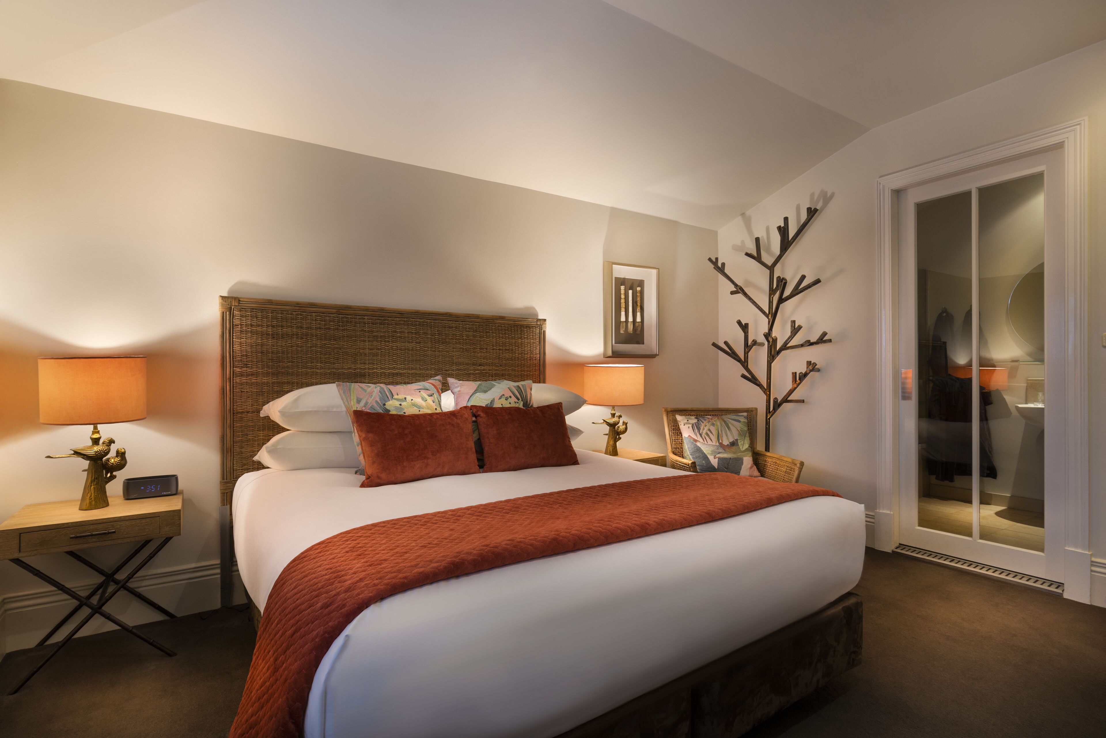 A hotel room with a large bed, two bedside tables, a nightstand, a wall-mounted clock, a framed picture, and a sliding glass door leading to a bathroom.