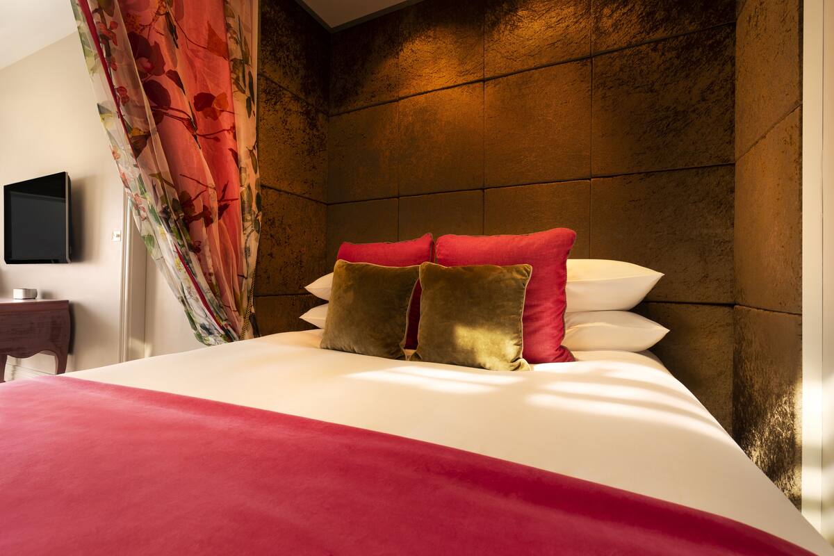 A neatly made bed with red and gold pillows, a television on a bedside table, and a curtain with a floral pattern.