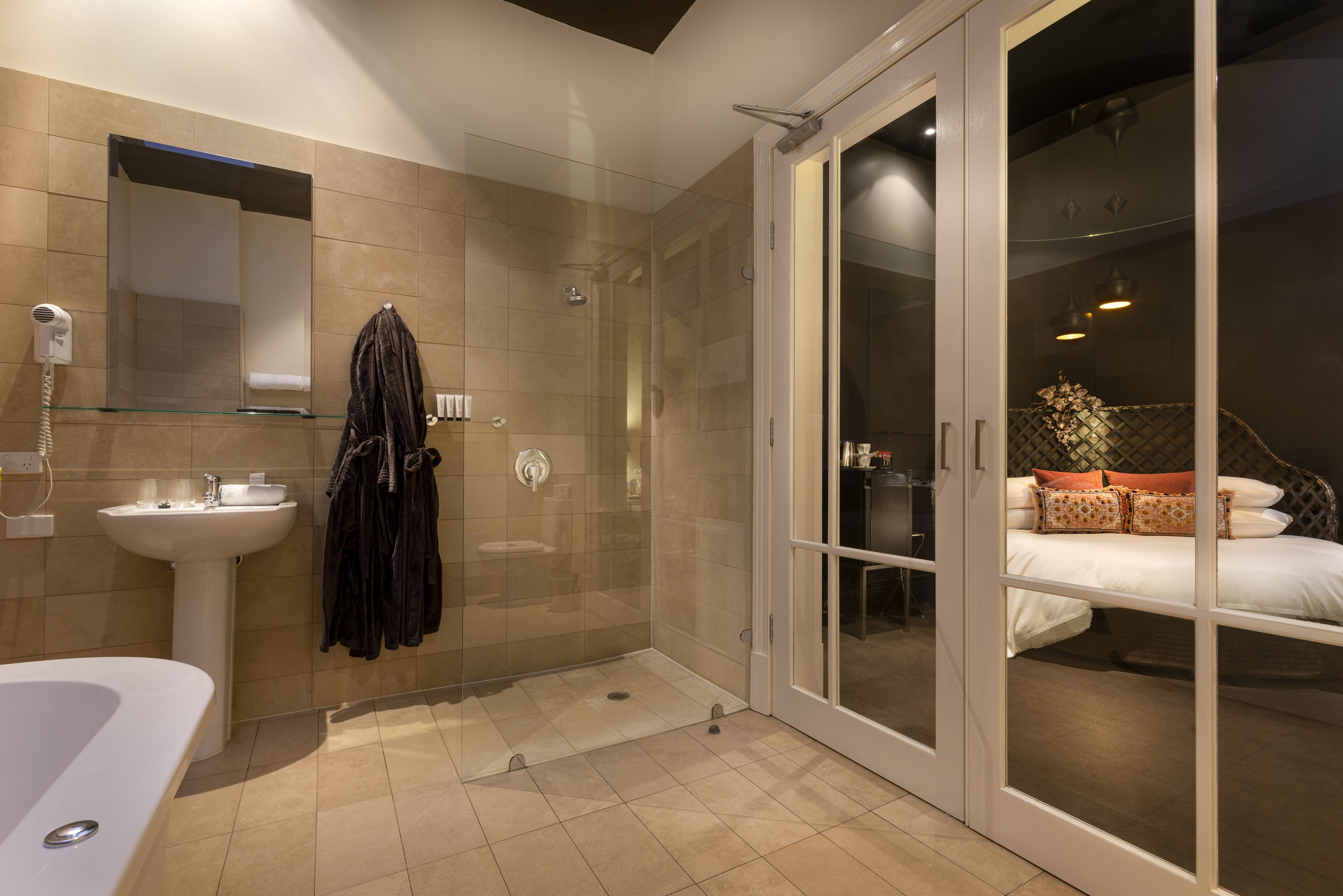 A modern bathroom with a glass shower enclosure, a white sink, and a glass shower door leading to a bedroom with a bed and decorative pillows.