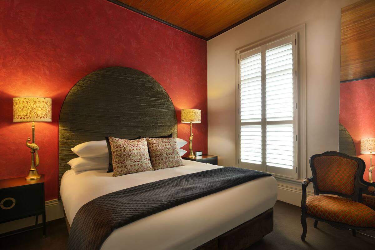 A bedroom with a large bed, two bedside tables with lamps, a chair, and a window with blinds.