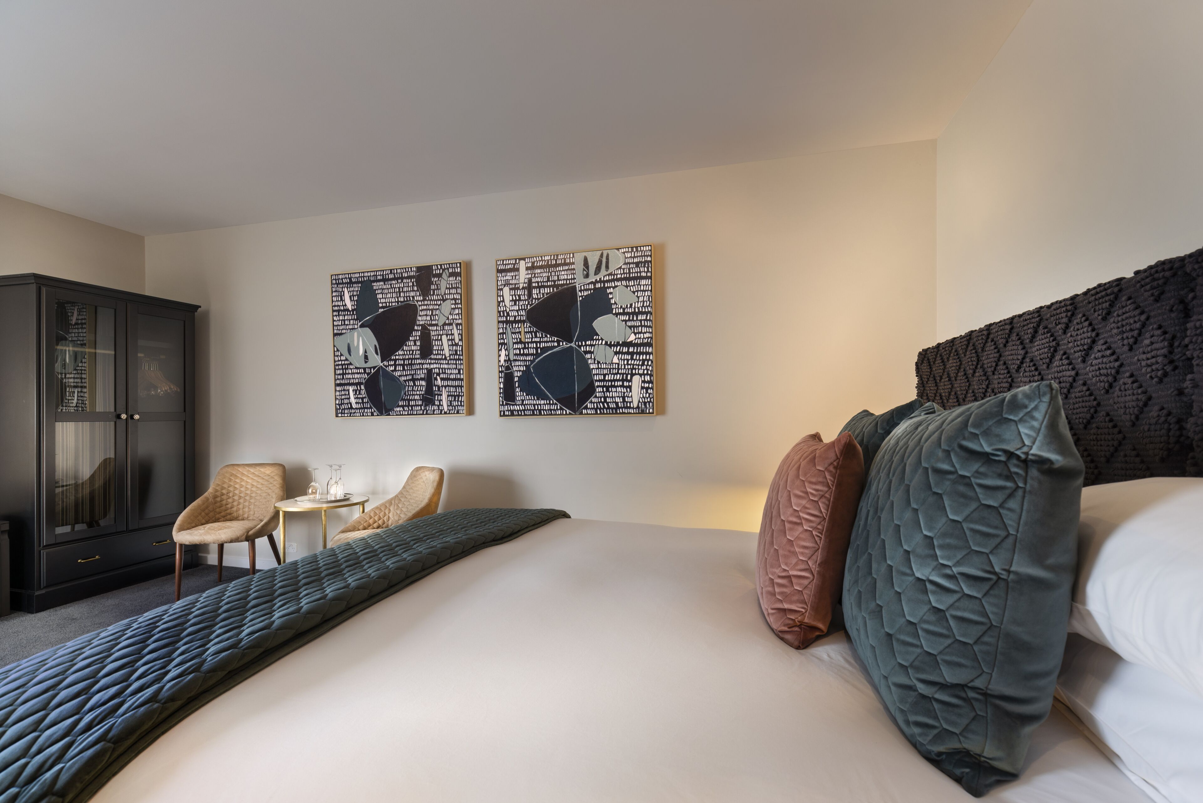 Ein Hotelzimmer mit einem großen Bett, zwei Wandbildern, einem dunklen Kopfteil und einer Sitzecke mit Stuhl und Tisch.