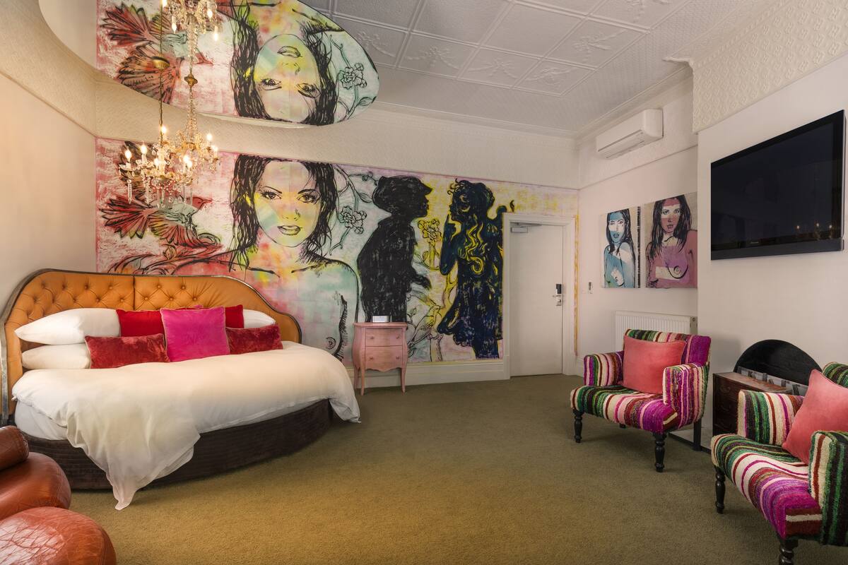 A bedroom with a large mural, a tufted headboard bed, a small bedside table, a TV, and striped armchairs.