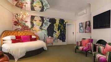 Une chambre à coucher avec une grande fresque murale, un lit avec une tête de lit capitonnée, une petite table de chevet, un téléviseur et des fauteuils à rayures.