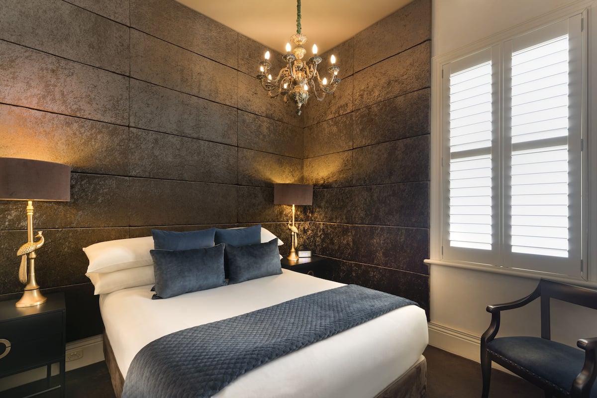 A bedroom with a large bed, two bedside tables with lamps, a chandelier, and a window with blinds.