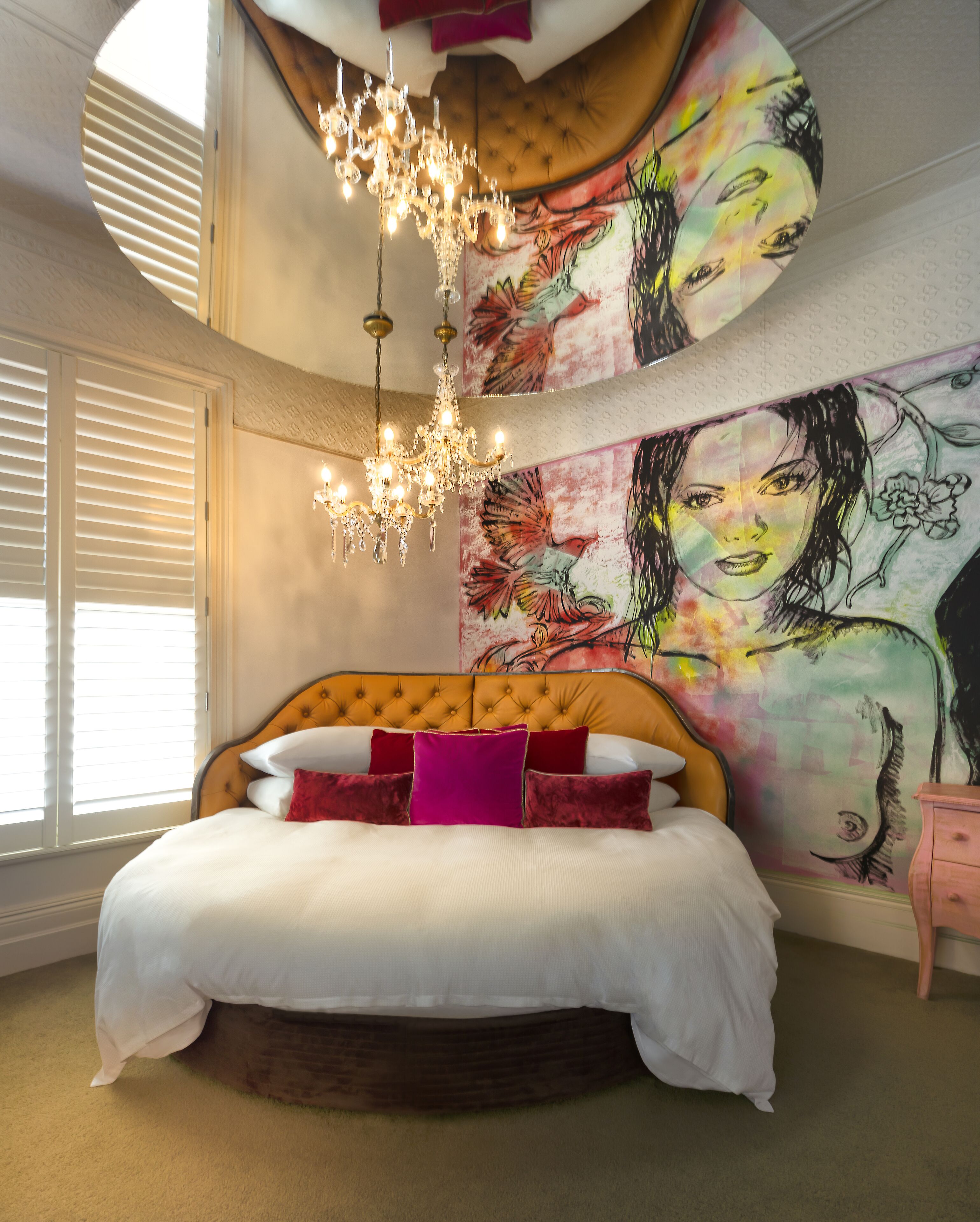 Ein Schlafzimmer mit einer gepolsterten Kopfteile, einem großen Bett mit weißer und roter Bettwäsche, einer Chandelieber und einem farbenfrohen Wandbild.