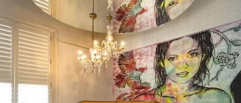 A bedroom with a tufted headboard, a large bed with white and red bedding, a chandelier, and a colorful mural on the wall.