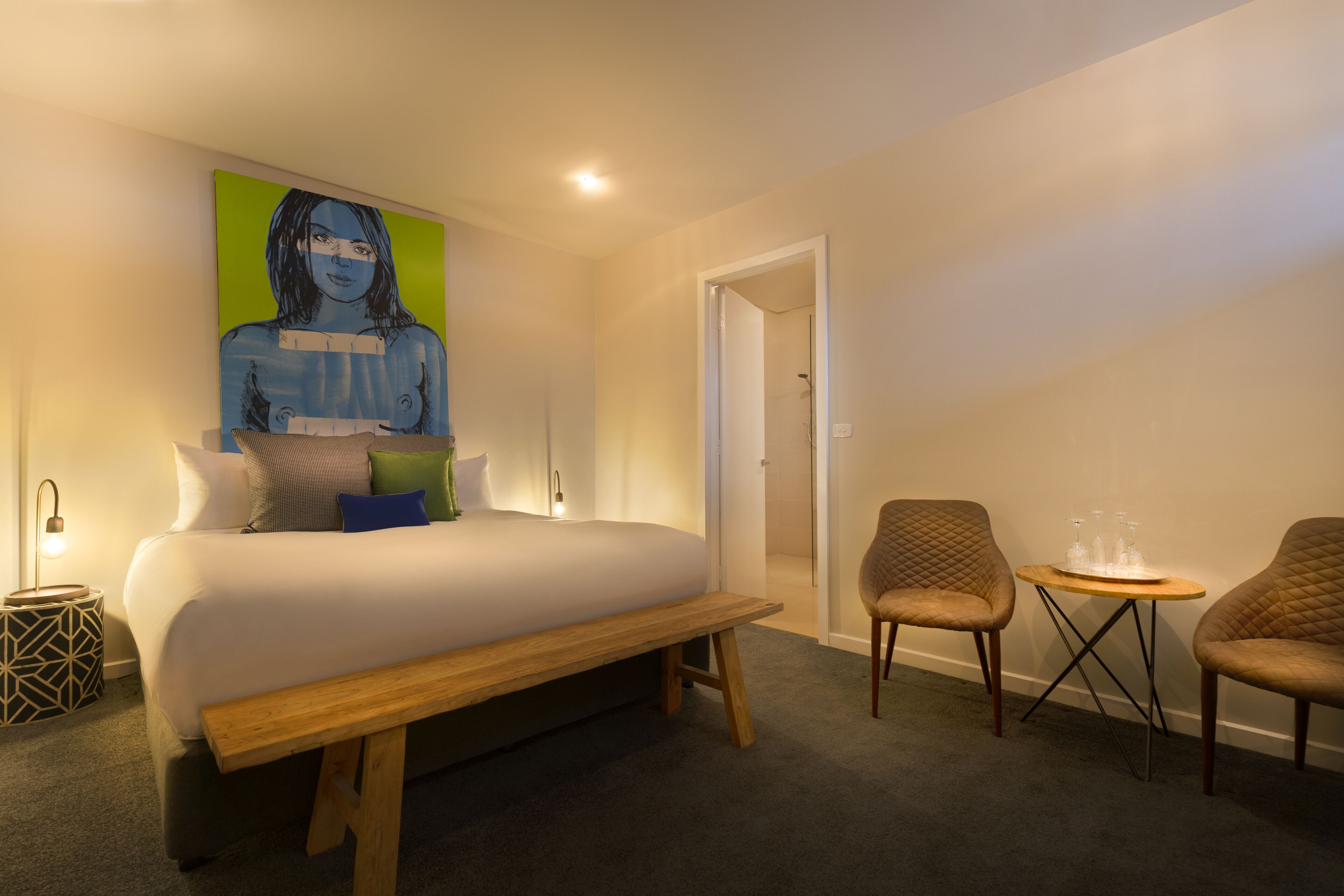 A modern hotel room with a large bed, a bench, two armchairs, a small table, and a painting on the wall.