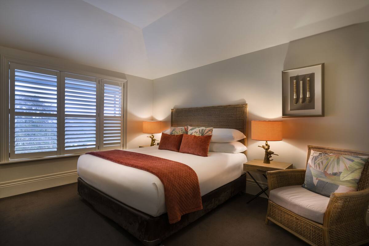 A neatly arranged bedroom with a large bed, two bedside lamps, a wicker chair, and a framed picture on the wall.