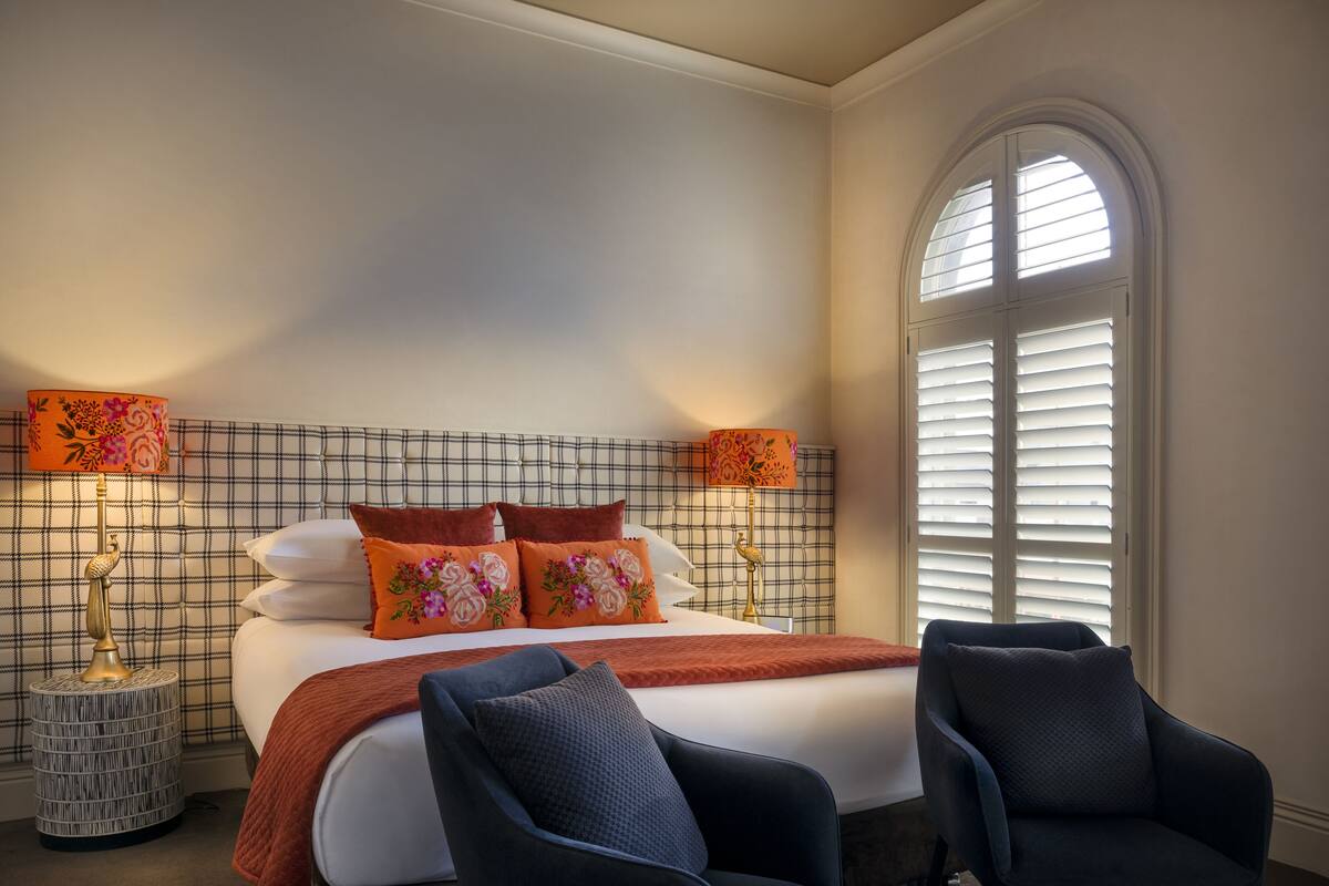 A hotel room with a bed, two chairs, a nightstand with a lamp, and a window with shutters.