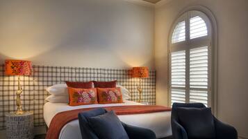 A hotel room with a bed, two chairs, a nightstand with a lamp, and a window with shutters.