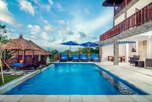Una piscina con sillas de descanso y sombrillas, una estructura de techo de paja y un edificio de varios niveles con terraza.