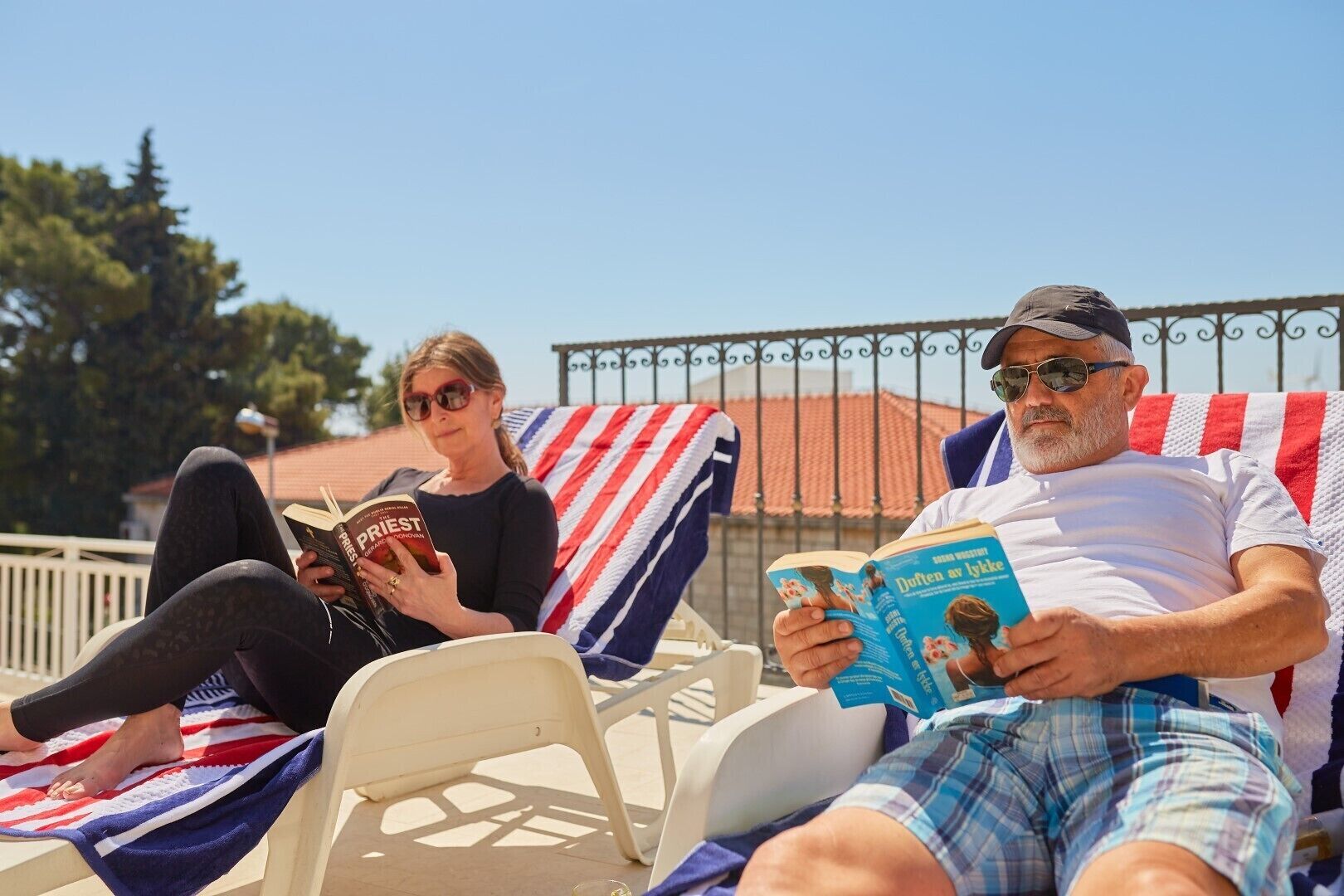 Two people relaxing on sun loungers with striped towels, reading books.