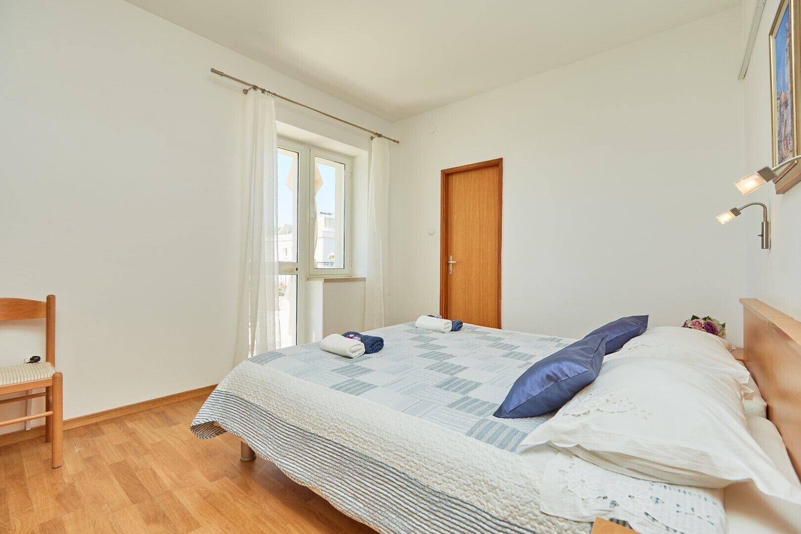 A bedroom with a bed, a wooden chair, a window with curtains, a door, and a wall-mounted lamp.
