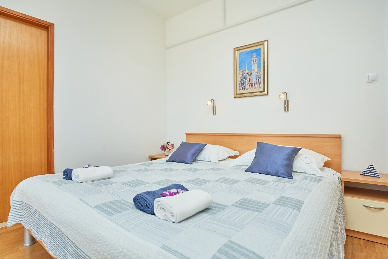 Ein ordentlich gemachtes Bett mit einer Steudecke, Kissen und aufgerollten Handtüchern. Ein Nachttisch mit Lampe und einem kleinen dekorativen Gegenstand steht daneben. Ein gerahmtes Bild hängt über dem Bett an der Wand.
