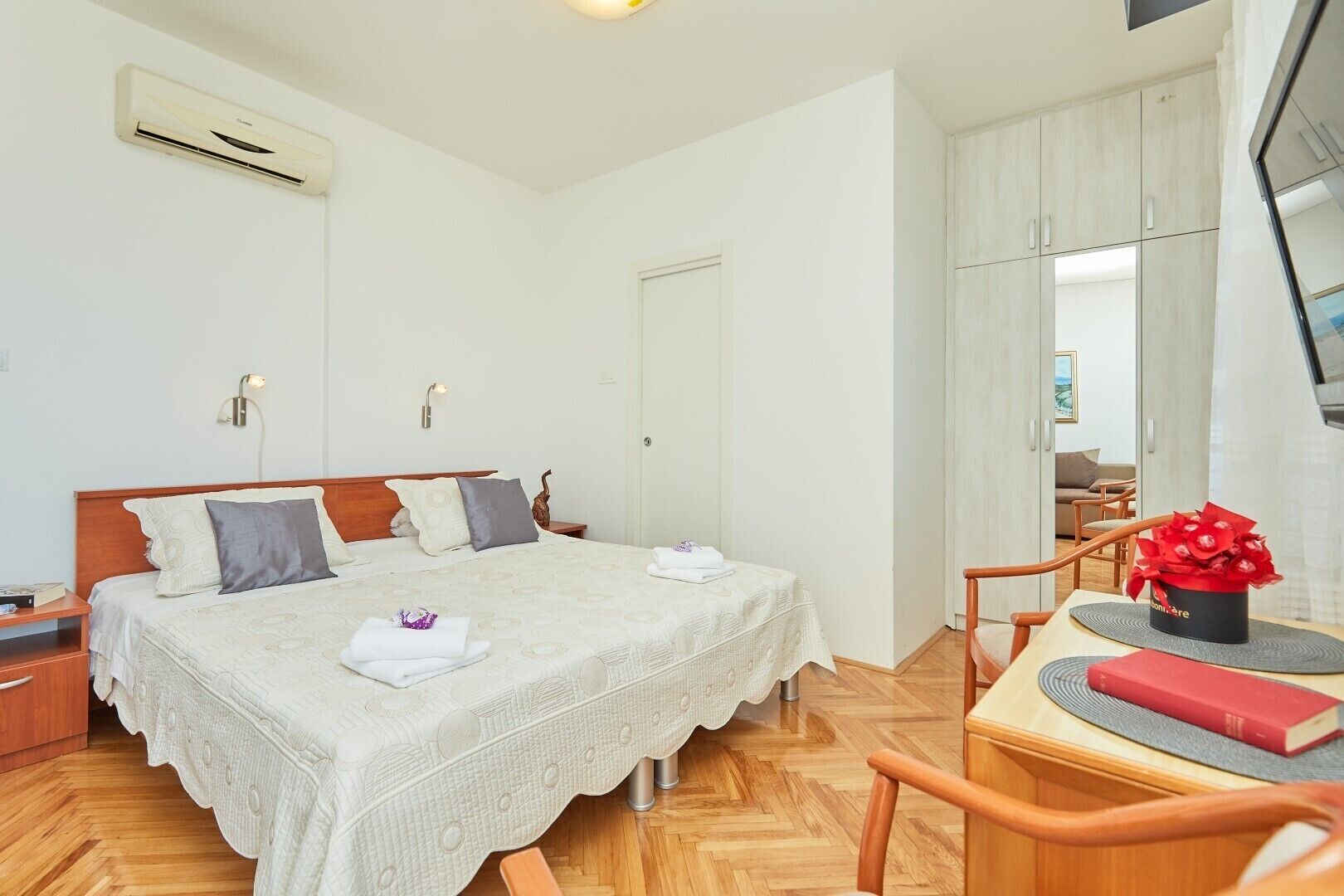 A bedroom with a bed, bedside tables, a desk with a chair, a TV, and an air conditioning unit.