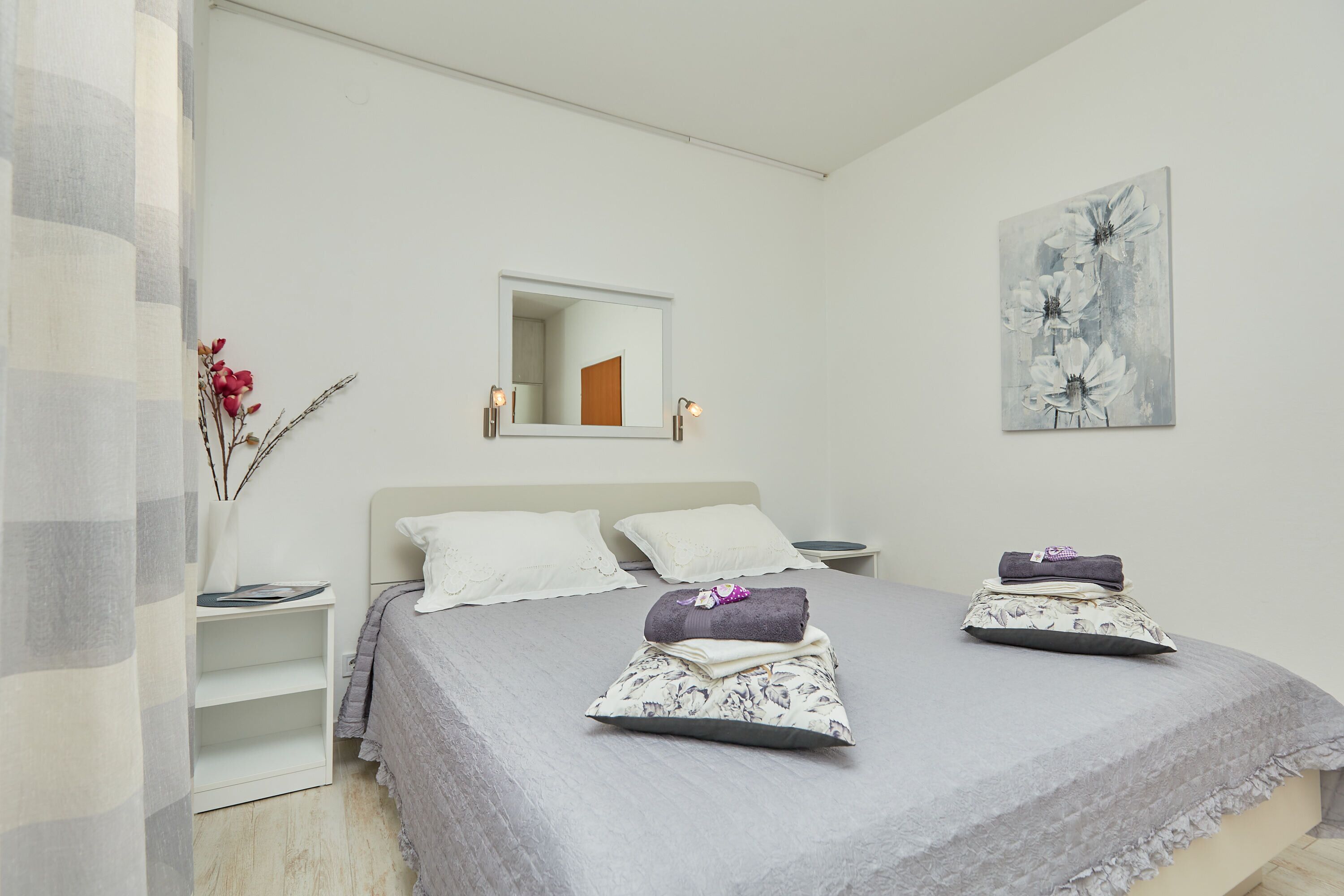 A neatly made bed with pillows and a folded blanket, a bedside table with a lamp, a wall-mounted mirror, and a floral painting.