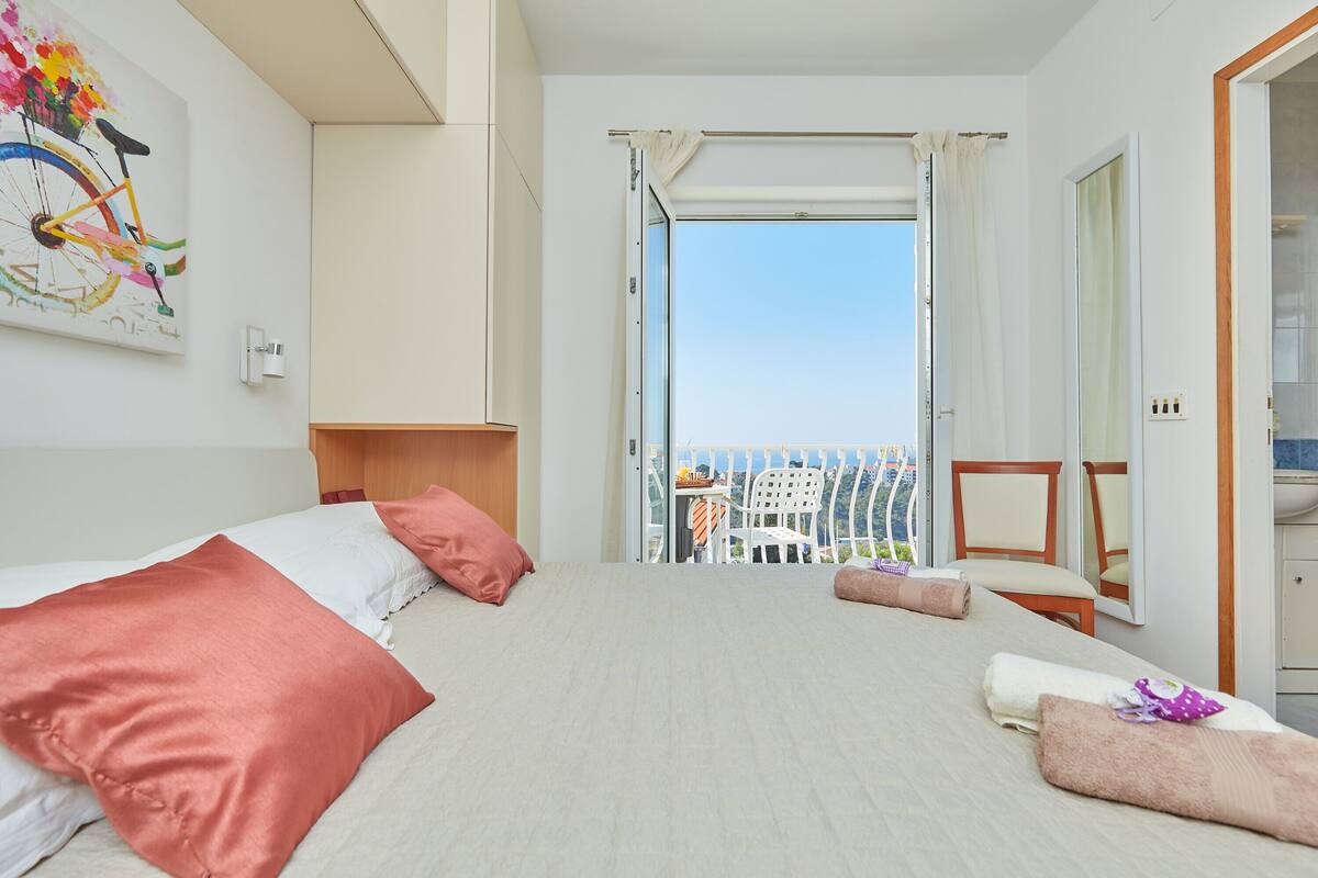 Een hotelkamer met een bed, uitzicht op zee en een balkon met stoelen.