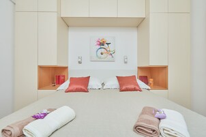 Moderni hotellihuone, jossa on suuri sänky, kaksi yöpöytää, seinälle ripustettu taideteos sekä puinen hylly, jossa on kirjoja ja kynttilä.