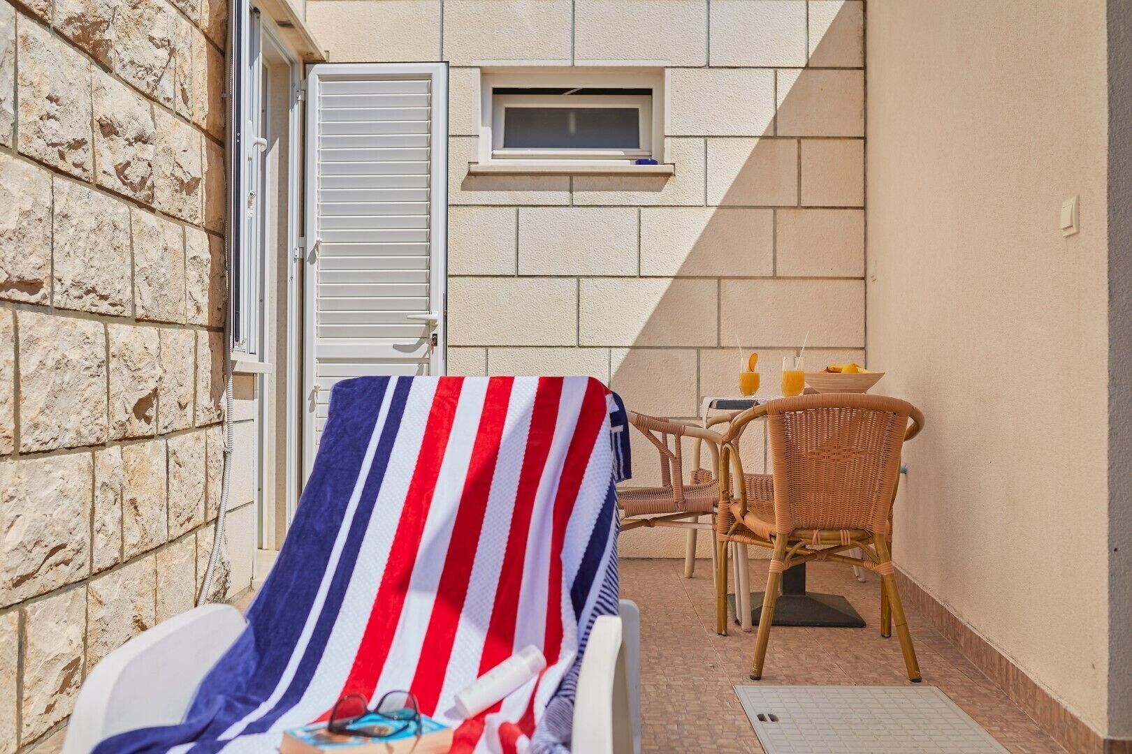 Een zonnig buitengebied met een gestreepte handdoek, rieten stoelen en een tafeltje met drankjes.