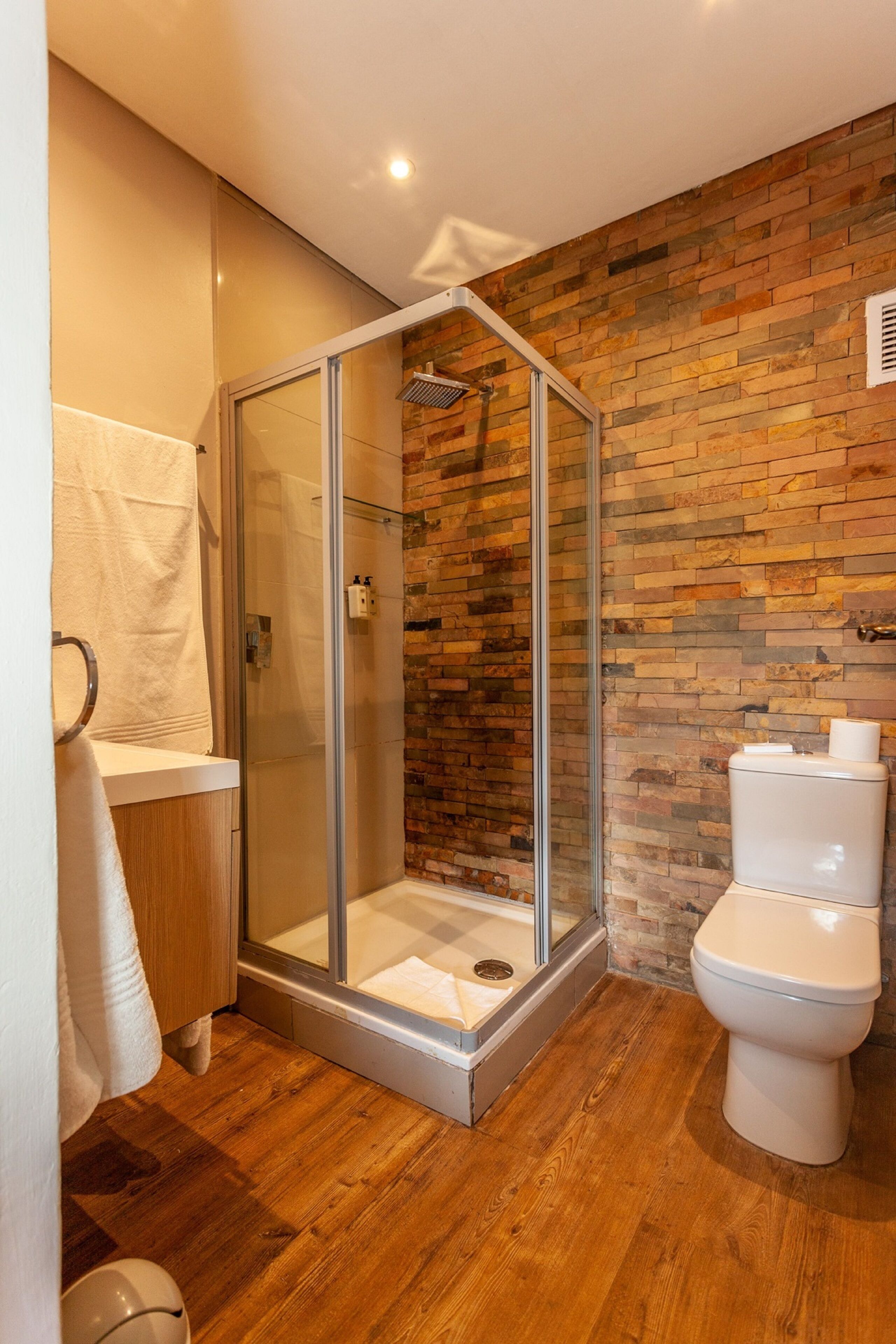 A modern bathroom with a glass-enclosed shower, wooden flooring, and a brick accent wall.