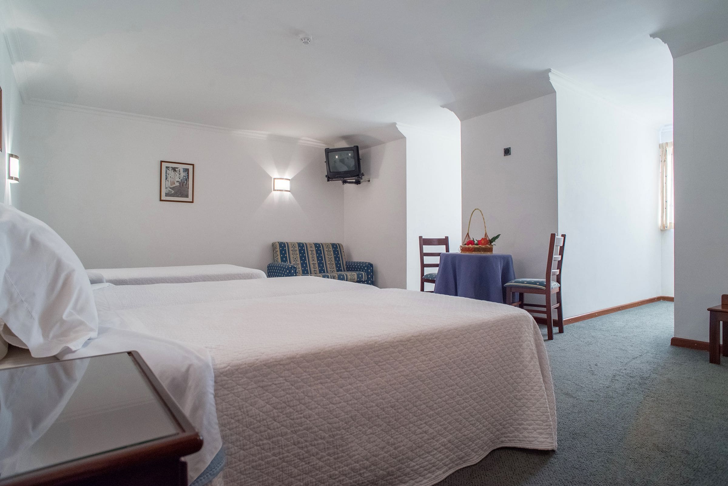 Quarto de hotel com duas camas, uma pequena mesa com uma cadeira, uma televisão fixada na parede e um quadro emoldurado na parede.
