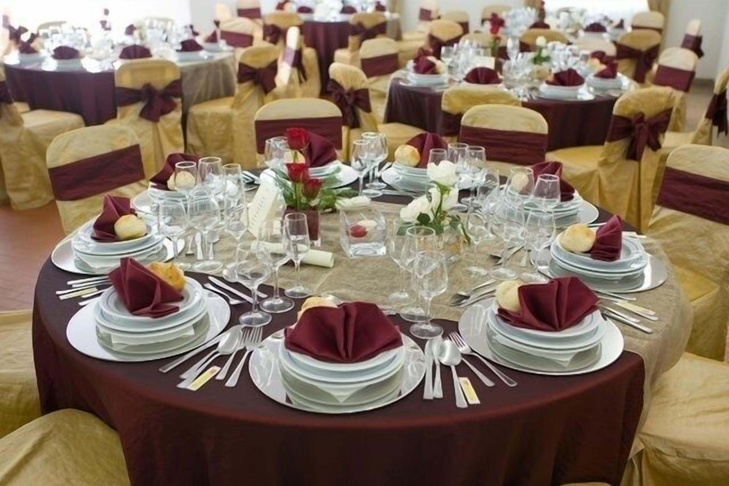 Un salón de banquetes con mesas redondas dispuestas para un evento formal, con platos blancos, vasos transparentes y servilletas rojas.