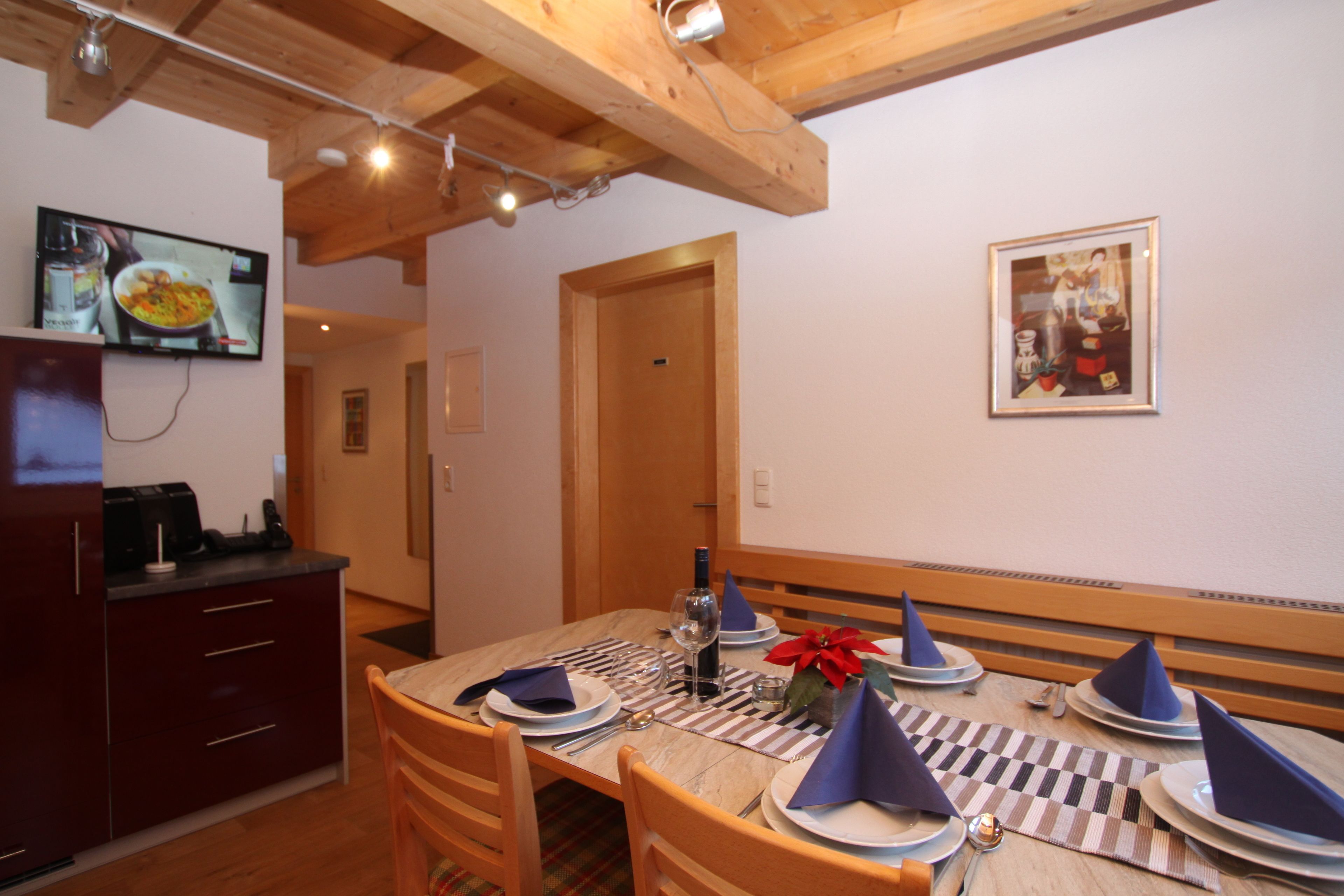 Un comedor con una mesa de madera dispuesta para cuatro, un televisor montado en la pared y una puerta que da a otra habitación.