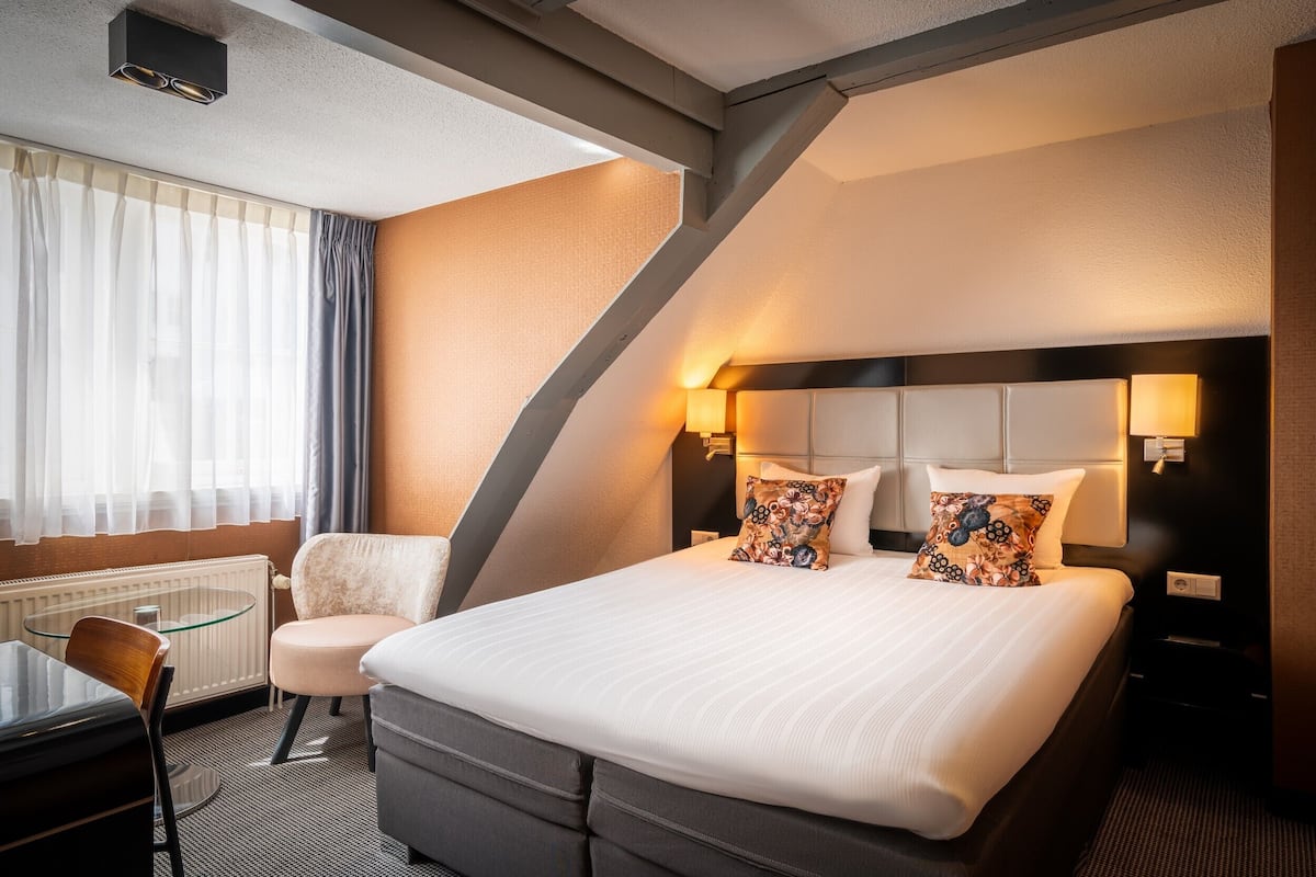 A hotel room with a large bed, a chair, a glass table, and a desk.