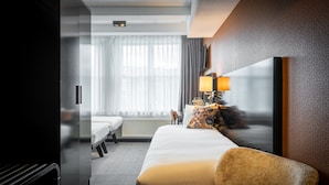 A hotel room with a bed, a chair, a lamp, a TV, and a window with curtains. - Hotel Mansion (Amsterdam)