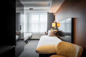 Une chambre d’hôtel avec un lit, une chaise, une lampe, une télévision et une fenêtre avec des rideaux.