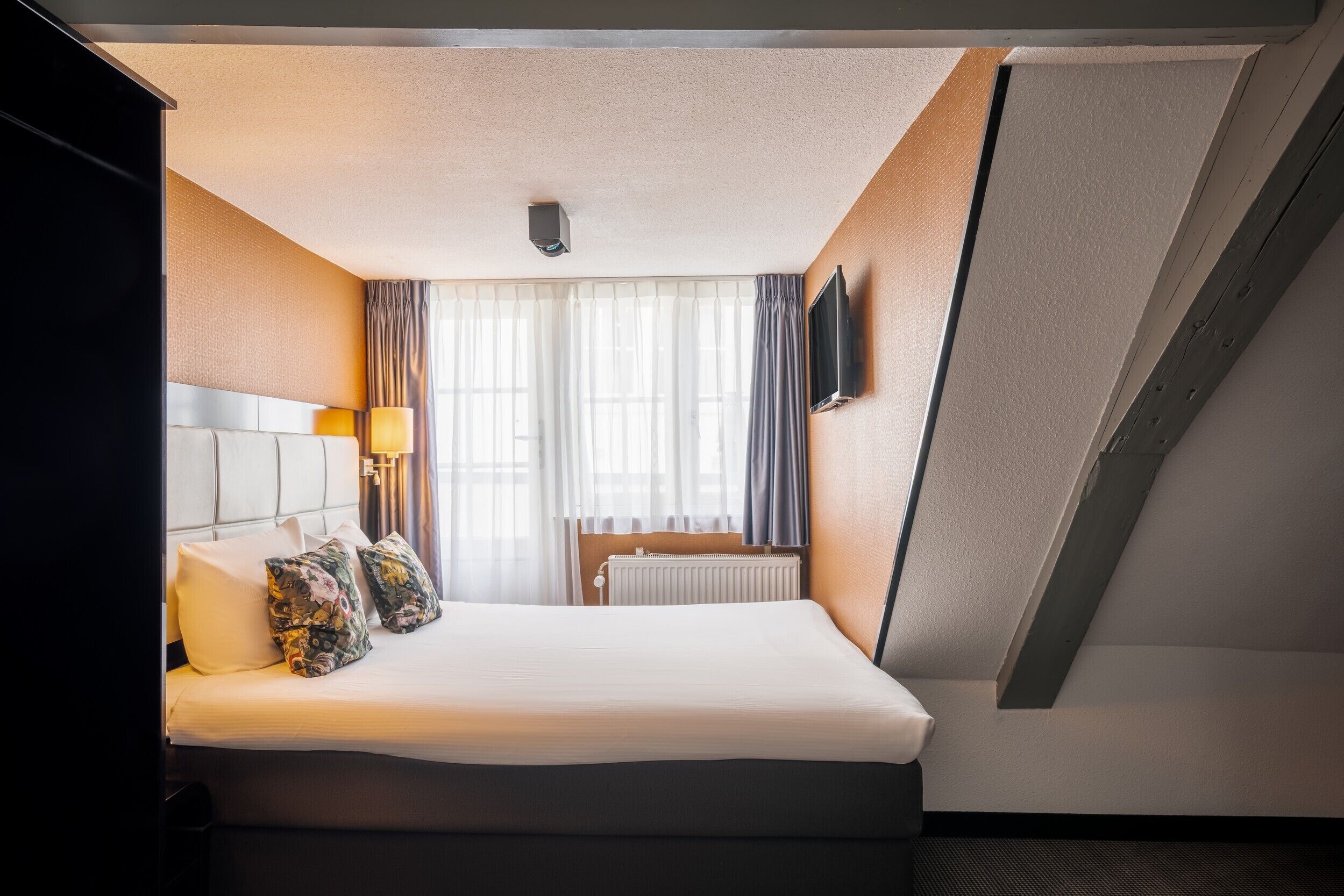 Habitación de hotel con cama, televisión de pantalla plana en la pared, ventana con cortinas y radiador.