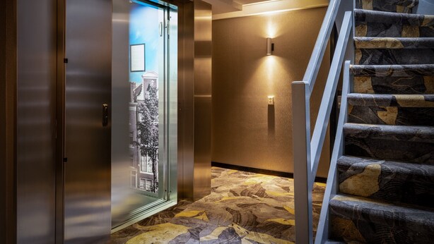 A modern elevator with a glass door, a carpeted floor, and a staircase with patterned steps. - Hotel Mansion (Amsterdam)