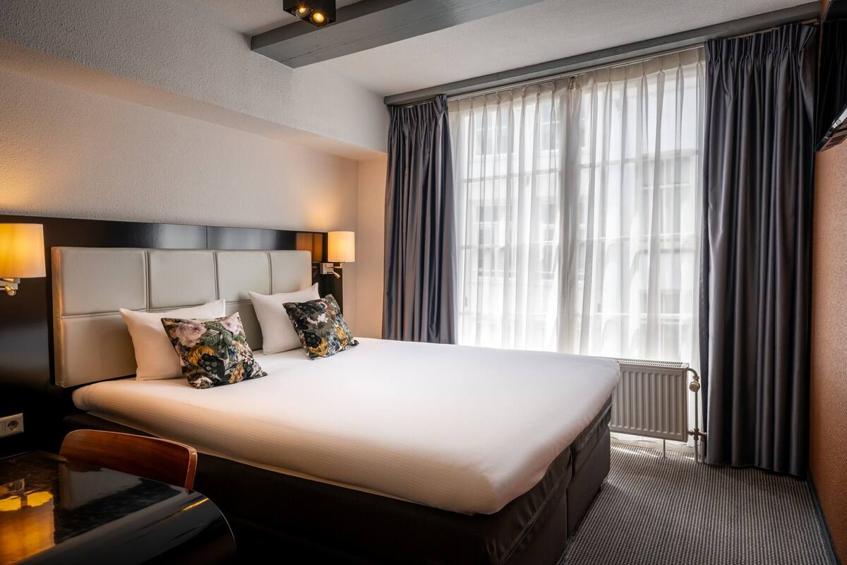 Een hotelkamer met een groot bed, nachtlampjes, een radiator en een raam met gordijnen.