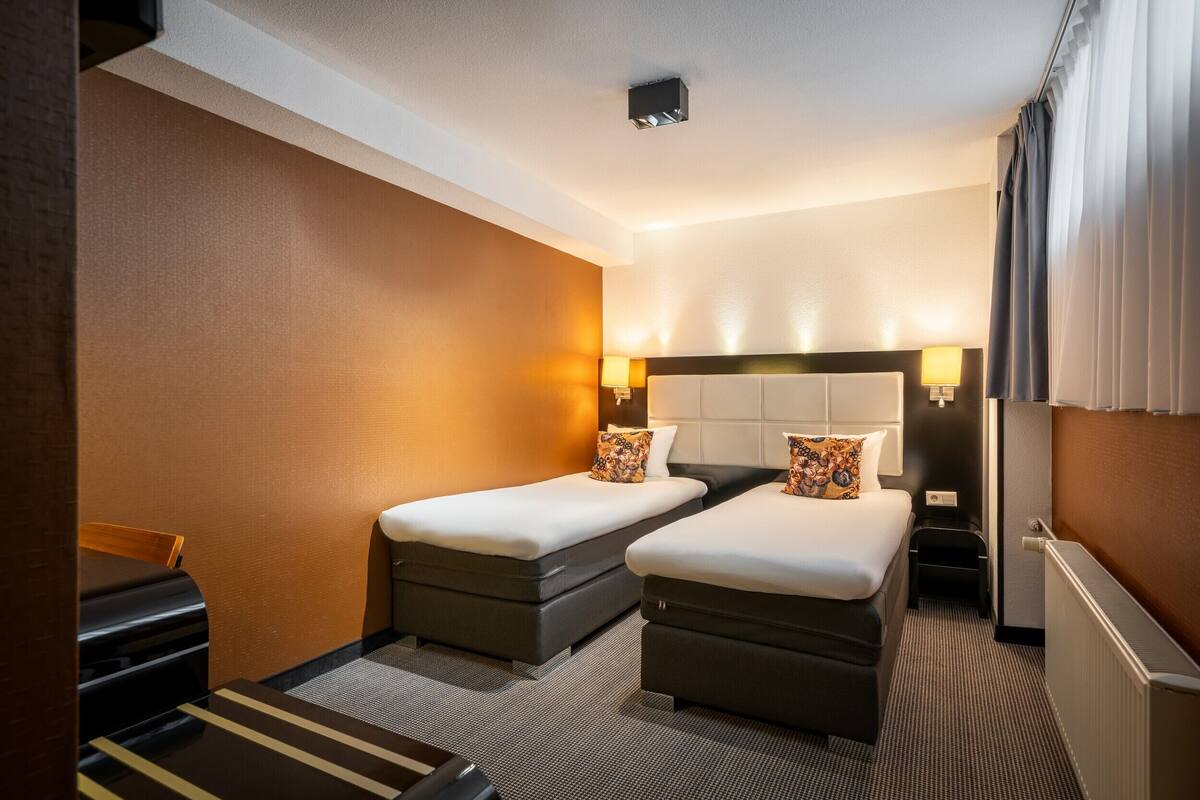 Een hotelkamer met twee bedden, een bureau, een stoel, een radiator en een raam met gordijnen.