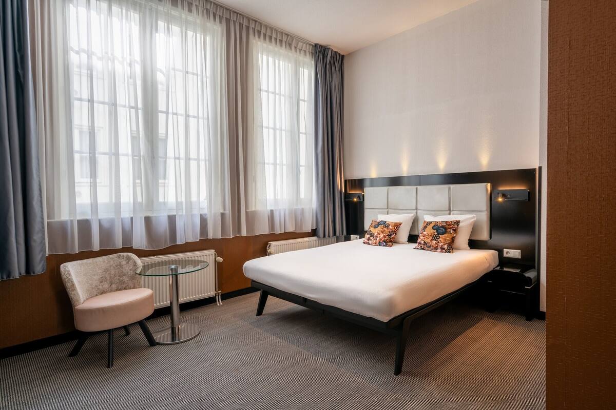 Een hotelkamer met een bed, een stoel, een tafeltje en grote ramen met gordijnen.