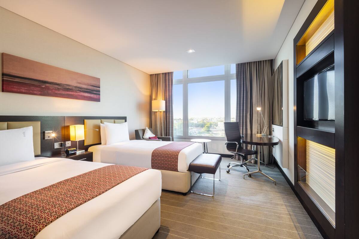 A hotel room with two beds, a desk, a chair, a TV, and a large window offering a city view.