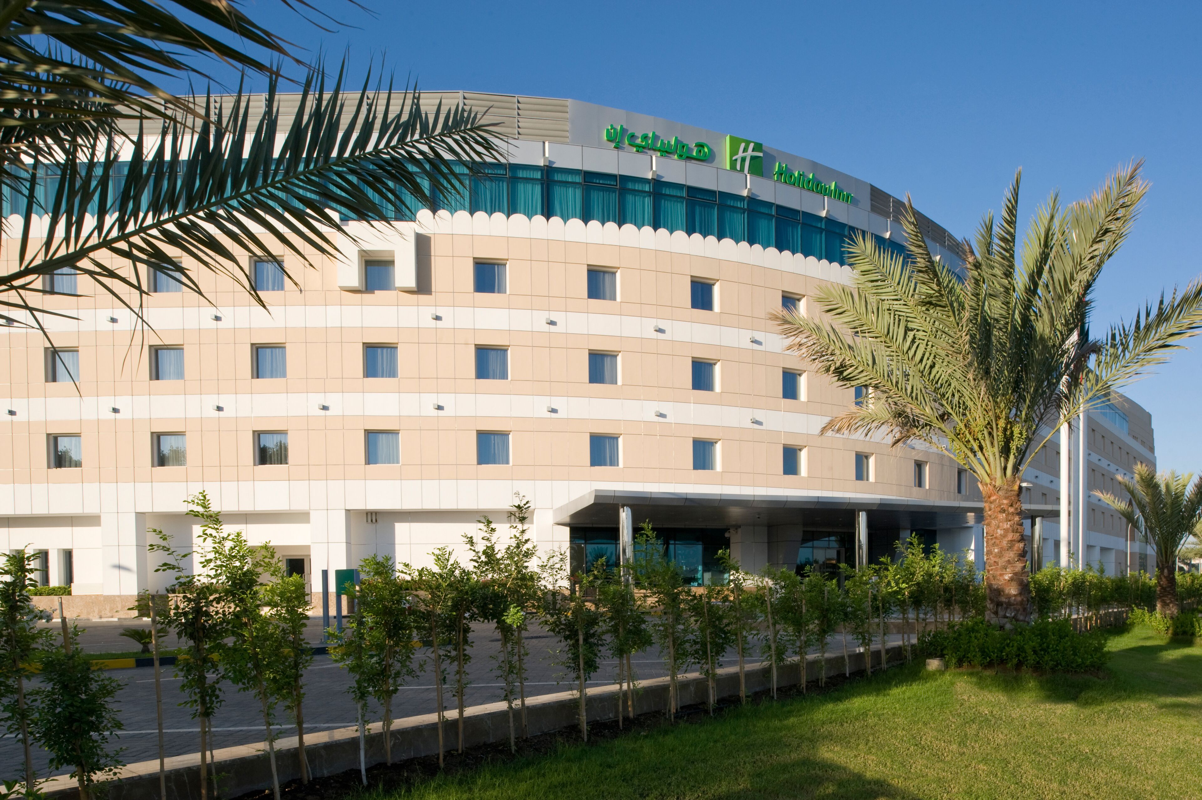 A modern hotel building with a curved facade, multiple windows, and a well-maintained lawn with young trees.
