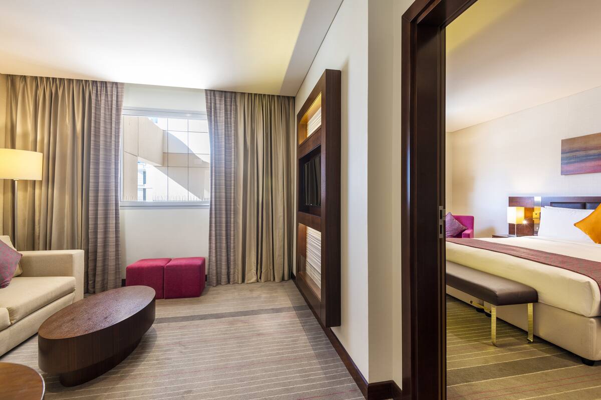 A hotel room with a sofa, a coffee table, a bed, a bench, and a window with curtains.
