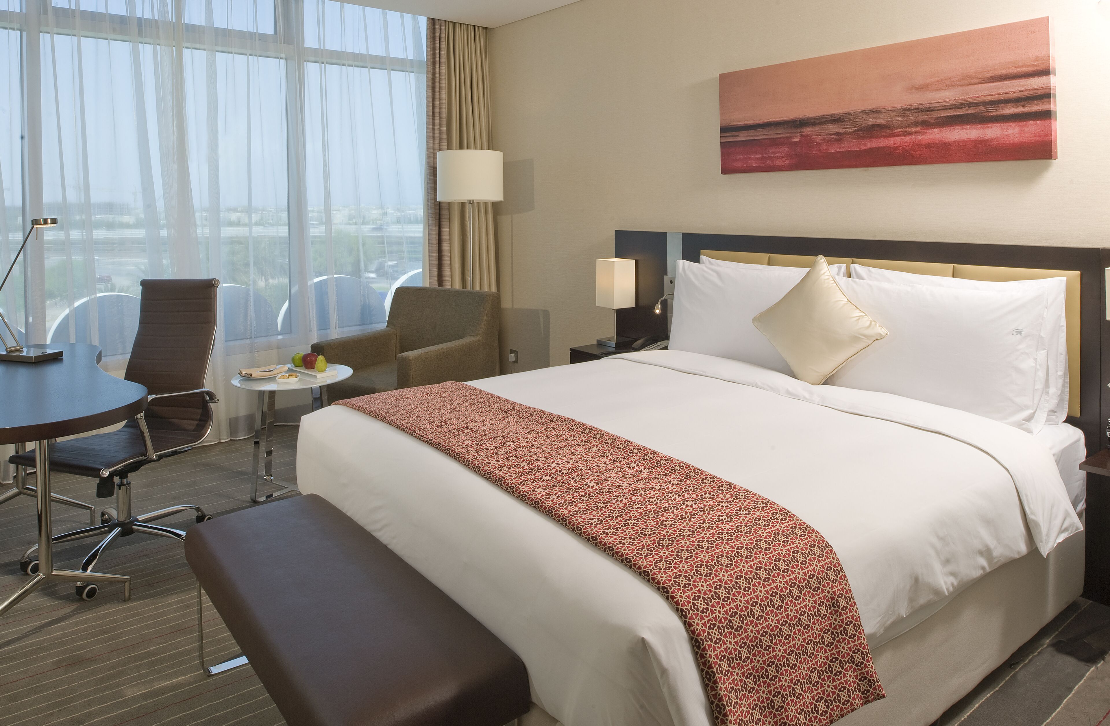 A hotel room with a large bed, a desk, a chair, a small round table, and a view of the city through large windows.